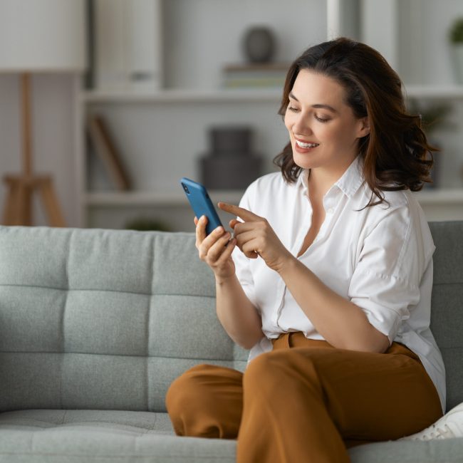 Happy casual beautiful woman is using a phone sitting on a sofa at home.